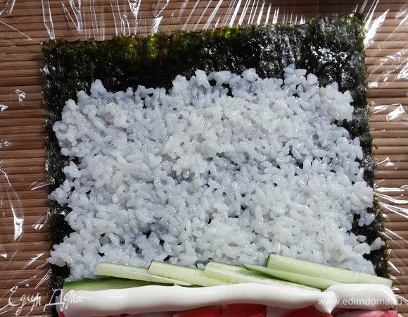 На лист выложить рис тонким слоем, немного не доходя до одного края. На другом краю выложить семгу, крабовые палочки, порезанные огурцы. Смазать майонезом. Посолить.