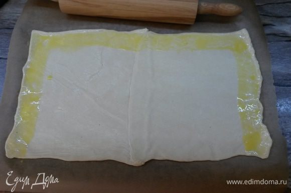 Размороженное тесто на присыпанном мукой столе раскатываем в прямоугольник 40х20 см, толщиной 4 мм. С помощью скалки перекладываем тесто на бумагу для выпечки. Смазываем 2 коротких и одну длинную стороны теста яйцом с молоком (как на фото). Ширина смазанных полос примерно 4 см.