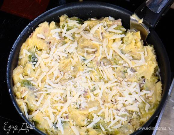 Дно сковороды, где жарился лук с грудинкой, посыпать половиной натертого сыра, выложить картофельно-капустную массу, разровнять ее и посыпать оставшимся сыром.