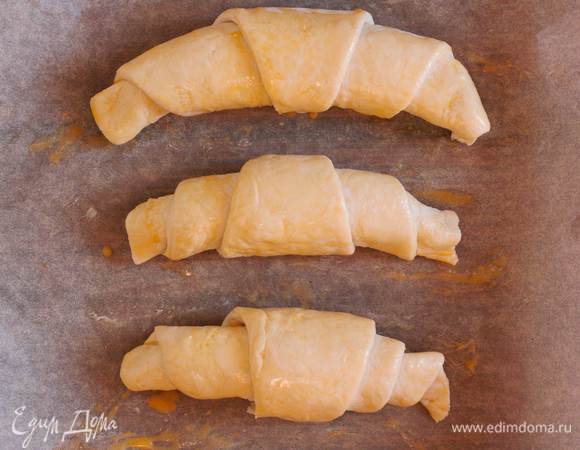 Тесто раскатайте на присыпанной мукой поверхности в прямоугольник толщиной 3 мм. Разрежьте его поперек, так чтоб получилось несколько прямоугольников. Далее каждый прямоугольник наискосок, на два треугольника. Каждый треугольник смажьте растопленным сливочным маслом. В основание выложите пару кубиков масла из морозилки и сверните тесто в рулеты. Получившиеся круассаны выложите на застеленный пергаментом противень и смажьте взбитым яйцом, и уберите в разогретую до 200°C духовку на 20-25 минут, или до появления румяной корочки.