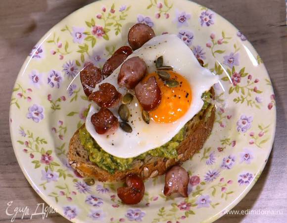 Подсушенный хлеб намазать массой из авокадо, сверху выложить глазунью, кусочки колбаски и посыпать все тыквенными семечками.