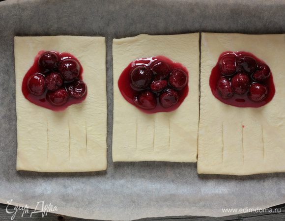 Зрительно разделите прямоугольники пополам и выложите на одну половину в центр по ложке вишни. На вторых половинах сделайте посередине по три надреза. Накройте начинку половиной листа с надрезами, защипните края.