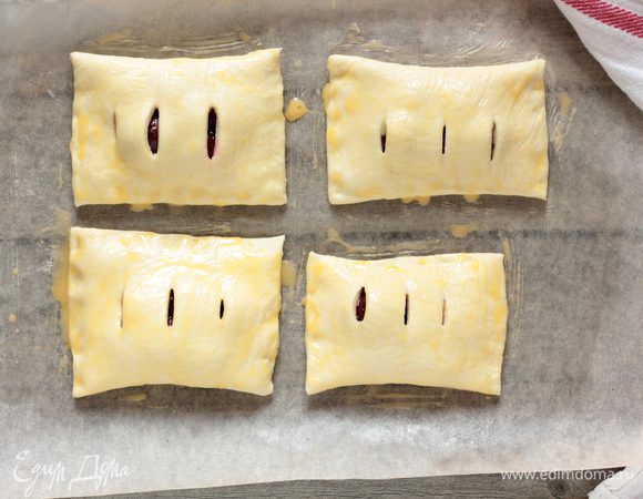 Противень выстелить бумагой для выпечки, смазать сливочным маслом и выложить вишневые слойки, смазать их взбитым с молоком яйцом. Посыпьте сахаром и уберите в разогретую до 190°C духовку на 20–25 минут или до появления румяной корочки.