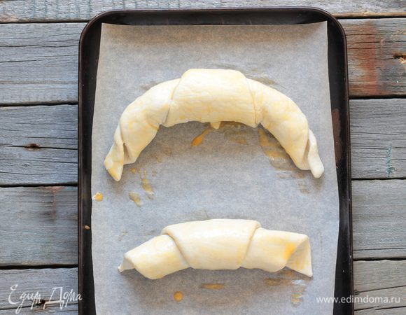 Каждый треугольник смажьте растопленным сливочным маслом. В основание выложите пару кубиков замороженного сливочного масла и сверните тесто в рулеты, двигаясь от основания треугольника к острому углу. Получившиеся круассаны переложите на застеленный пергаментом противень и смажьте взбитым яйцом, и уберите в разогретую до 200°C духовку на 20-25 минут или до появления румяной корочки.