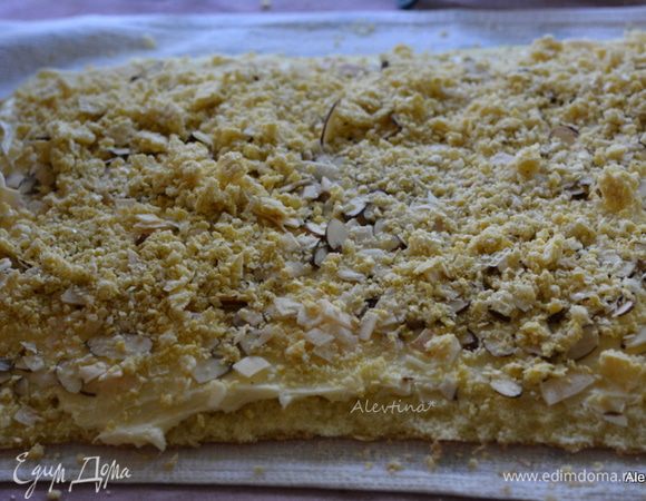 Развернуть рулет. Смазать частью приготовленной начинки. Выложить сверху вафельную крошку, миндальные лепестки, кокосовую стружку.