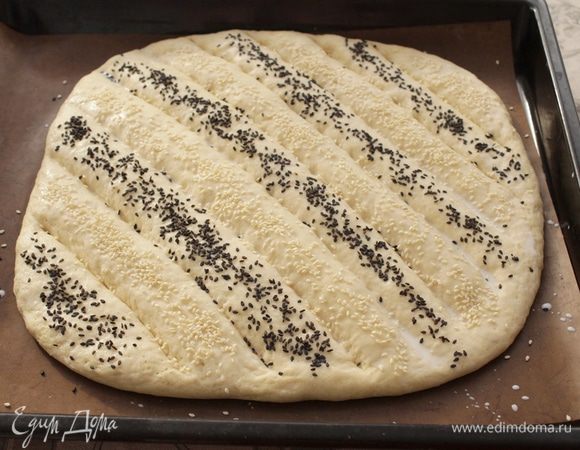Выложить тесто на противень, застеленный бумагой. Руками аккуратно растянуть тесто в лепешку, толщиной 1-1,5 см. Накрыть полотенцем и оставить, чтобы подошло, минут на 40.
Когда тесто поднимется, острым ножом нанести разрезы, смазать лепешку молоком, посыпать кунжутом.