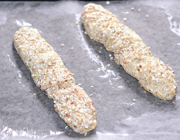 На рабочую поверхность высыпать овсяные хлопья. Руки смазать оливковым маслом (тесто немного липкое), вынуть тесто из хлебопечки. Положить его на хлопья и растянуть руками. Можно сразу разделить тесто на 2 части, а можно делать 1 багет. Затем, взявшись за концы заготовки, перекрутить несколько раз.

