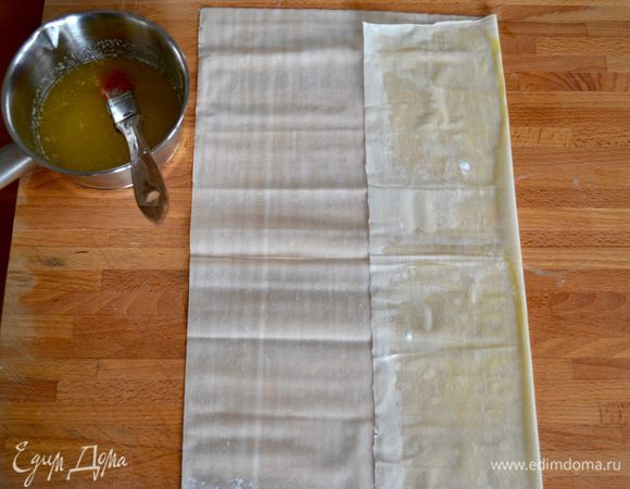 Растопите сливочное масло (его может понадобиться и чуть больше, чем указано в ингредиентах) и вместе с кисточкой приготовьте его рядом с тем местом, где будете работать с тестом.
Подготовьте тесто фило. Достаньте из упаковки, разверните готовые листы, накройте их пищевой пленкой и сверху кухонным полотенцем. Работать с тестом будем очень быстро, т.к. оно начинает сохнуть и рваться. Положите перед собой один лист теста фило. Визуально поделите его на три полоски. Правую (перед собой) полоску теста смажьте с помощью кисточки маслом. И заверните в центр, сложив лист теста на 1/3. Также смажьте маслом крайнюю левую полоску и еще раз заверните тесто...