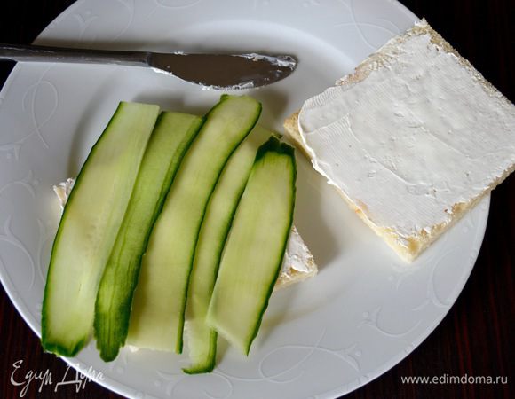 Начинаем "собирать" сэндвич... На одну сторону каждого ломтика хлеба намазать тонким ровным слоем сыр Филадельфия. Сверху выложить наискосок полоски огурца. Аккуратно придавить и при необходимости опять промокнуть бумажной салфеткой (бумажным полотенцем).