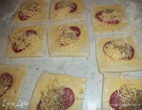 Колбасу нарезать тоненькими кружочками. Голландский сыр натереть на терке со средними отверстиями.
На каждый квадратик теста выложить по кружочку колбасы, присыпать натертым сыром. 
Сверху слегка присыпать орегано.
