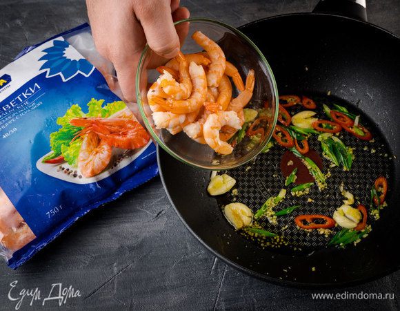 Разогреть в сковороде кунжутное и оливковое масло и обжарить зеленый лук и чили. Чеснок почистить, раздавить ножом и добавить в сковороду. Имбирь почистить, натереть на мелкой терке (должно получиться 1/2 ч. ложки), отправить в сковороду с луком и чесноком, все перемешать. Добавить креветки и обжарить их вместе с овощами почти до готовности.
