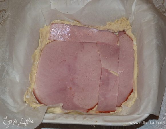 Жаропрочную форму застелить пекарской бумагой. Тесто выложить на дно, немного завернув бортики. Часть теста оставить для формирования бортиков. Выложить на тесто полоски ветчины, часть оставить для украшения.