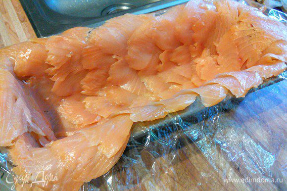 Прямоугольную форму для террина (~ 1,25 л объемом) слегка смазать маслом и выложить пищевой пленкой с запасом. Дно и стенки выложить лососем тонкой нарезки в нахлест (выбрать самые симпатичные кусочки), так же оставляя запас лосося, чтобы затем прикрыть террин. Если у вас не получилось оставить достаточно длинные свисающие кусочки лосося, чтобы затем прикрыть террин, то отложите несколько ломтиков рыбки из общего количества.