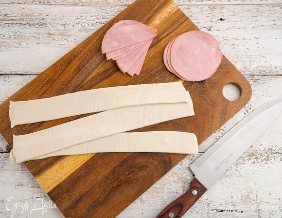 Слоеное тесто немного раскатываем и режем на полоски приблизительно 3 см. Колбасу режем тонкими кружочками и перерезаем пополам. Далее колбасу выкладываем так, чтобы было видно кружочки сверху полосок, а снизу ничего не торчало. И скручиваем туго колбасу с тестом. 