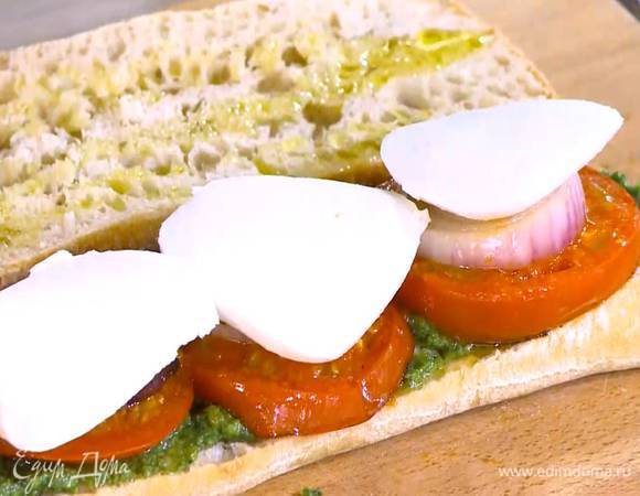 Чиабатту разрезать вдоль пополам и сбрызнуть оставшимся оливковым маслом, одну половину смазать соусом песто, выложить на нее обжаренные помидоры, лук и моцареллу.