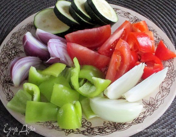 Подготовьте овощи для запекания. У перцев удалите семечки, а мякоть нарежьте на прямоугольные кусочки примерно 2х3 см. Помидоры нарежьте крупными дольками. Лук (тут половинка обычного репчатого и половинка сладкого фиолетового) разрежьте на 6 — 8 частей. Молодой кабачок или цукини — тонкими кружочками или ломтиками.
