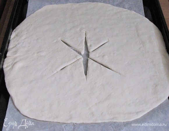 Подошедшее тесто обмять, снова сформировать шар и раскатать в круг. Круг с помощью скалки перенести на противень, застеленный пекарской бумагой. В центре круга сделать крестообразные разрезы.