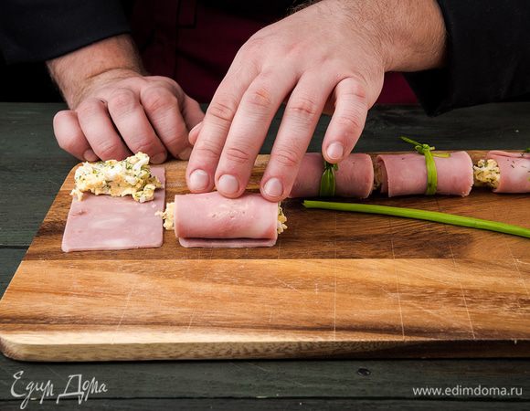 Ветчину нарезать тонкими слайсами по длинной стороне. На край каждого кусочка ветчины кладем начинку и сворачиваем рулетик. Стрелкой зеленого лука перевязать свиток.
