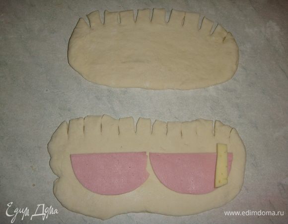 Колбасу тонко нарезать кружочками и пополам. Сыр нарезать брусочками. Каждый шарик раскатать в прямоугольник, сделать надрезы ножом с одной стороны (1 см). Положить кусочек колбасы. С одного края положить 1 брусочек сыра. Начать сворачивать тесто в рулетик с той стороны, где лежит сыр.