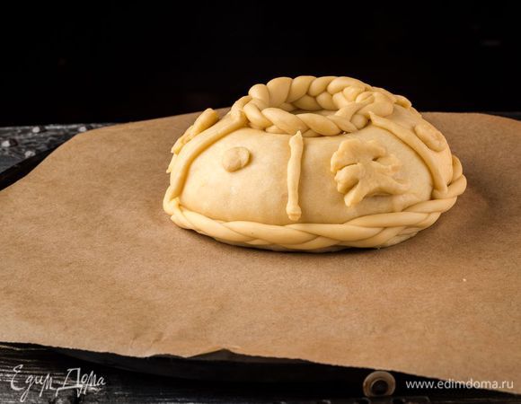 Рабочую поверхность присыпать мукой, выложить тесто и месить его 20 — 30 минут, чтобы оно стало гладким и не липло к рукам. Духовку нагреть до 200°С. От теста отрезать 100 г, из остатка сформировать шар и положить на противень, застеленный пергаментом. Шар смазать. Из отрезанного теста слепить украшения (цветы, косы, решетки, листья) и расположить их на каравае. Периодически нужно смазывать водой поверхность теста, чтобы не образовалась корка.