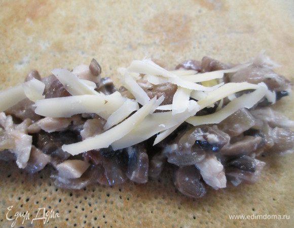 Кладем блин обжаренной стороной вверх и выкладываем на один его край начинку. Сверху посыпаем тертым сыром (используем 1/2 часть от общего количества). Сворачиваем рулетом.