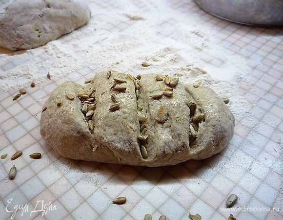 Подошедшее тесто разделяем на две части, формируем хлебушек, обваливаем в муке, перекладываем на противень, застеленный пекарской бумагой, делаем надрезы и посыпаем подсолнечными семечками. Оставляем заготовки подходить в тепле 40 минут, накрыв полотенцем.