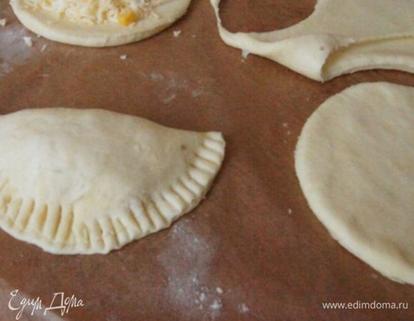 Раскатать тесто толщиной около 2 мм, вырезать круглые лепешки, поместить сырную начинку и немного кукурузы на каждый пирожок. Края смазать водой, чтобы они лучше склеились. Защипнуть пирожок, края придавить вилкой. Приклеить нужно хорошо, чтобы начинка не вытекла.