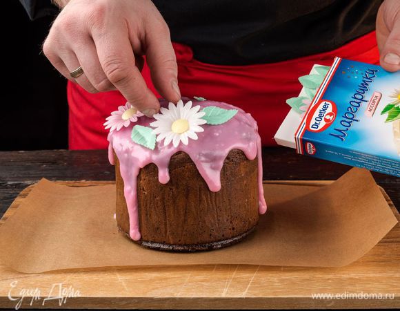 Срежьте уголок упаковки, покройте кулич глазурью и пока она не остыла, декорируйте кулич розовыми, белыми вафельными маргаритками и зелеными лепестками.