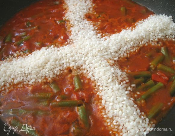 В сковороду налить 1 стакан бульона, перемешать. Очередь риса. Количество риса обычно определяют на глазок. Не новичок, а знаток и профессионал своего дела не измеряет объем риса, а высыпает его в бульон крестообразной горкой. Главное правило, которого следует придерживаться при приготовлении паэльи — рис должен возвышаться над поверхностью на 1,5-2 см, не выше. Бульон, перед тем как выкладывается рис, должен кипеть. Разровнять рис деревянной ложкой по поверхности, добавить бульон (2 стакана).