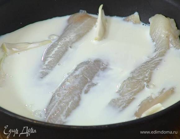Уложить в тяжелую сковороду куски размороженной рыбы кожей вниз, между ними поместить зубчики чеснока в кожуре, залить все молоком, затем влить немного воды, так чтобы пикша была полностью покрыта, и готовить под крышкой минут 10 до готовности.