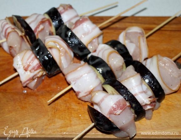 Деревянные шпажки предварительно замочить на 30 минут в воде. Кусочки курицы обернуть полосками бекона и чередуя с баклажаном нанизать на шпажки.