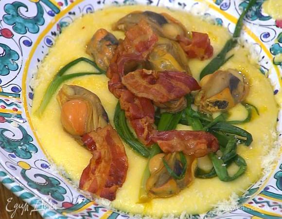 В готовую поленту добавить сливочное масло, большую часть натертого сыра, все вымешать и выложить на тарелку, сверху разложить зеленый лук и мидии, посыпать оставшимся пармезаном и украсить беконом.