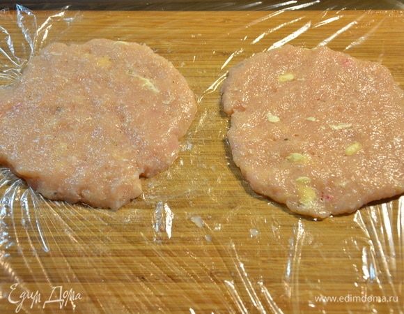 На разделочной доске расстелить пищевую пленку. Смазать растительным маслом. Намочить руки в холодной воде и слепить лепешку из куриного фарша. Выложить куриную лепешку на пленку и немного расплющить.