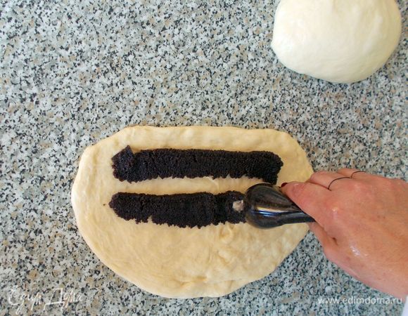 Готовое тесто делим пополам. Каждый «колобочек» (кусочек) теста раздавливаем и растягиваем на столе, смазанном растительным маслом, руками (не скалкой!).  Толщина должна получится примерно 1 см. Затем берем маковую начинку. Я использую уже готовую маковую начинку. Это намного упрощает и ускоряет процесс приготовления рулета. Готовая начинка по форме напоминает небольшую «колбаску». Делаем небольшой надрез (примерно 1–1,5 см) у основания маковой «колбаски» и выдавливаем из нее маковую начинку сразу на подготовленное тесто. Количество начинки — примерно половина «колбаски» на один рулет.