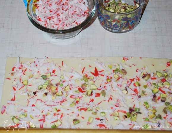 Тесто смазываем яйцом, посыпаем фисташками и крабовыми палочками, придавливаем скалкой.