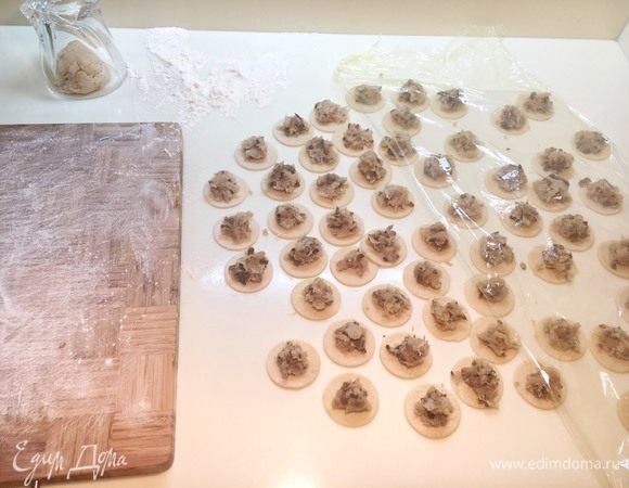 Выложите начинку на тесто, примерно 2/3 чайной ложки на пельмень. Накройте большую часть заготовки пленкой, чтобы тесто не заветрелись во время лепки. Присыпьте мукой дощечку или тарелку на которую поместите готовые пельмени, чтобы они не прилипли.