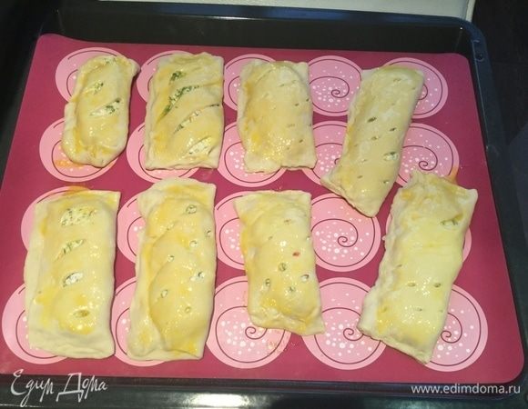 Выложите пирожки на противень, застеленный бумагой для выпечки или силиконовым ковриком. Слегка взбейте желток и смажьте им слойки.
