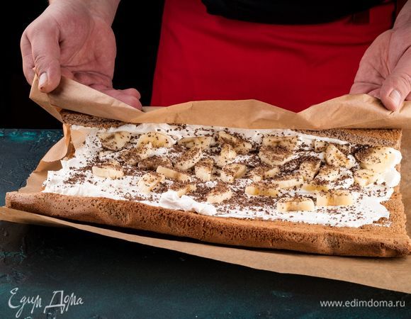 Смажьте поверхность бисквита взбитыми сливками. Сверху выложите полоски кружочков банана. А между полосками банана посыпьте шоколадом. Сверните рулет и уложите на тарелку швом вниз. 