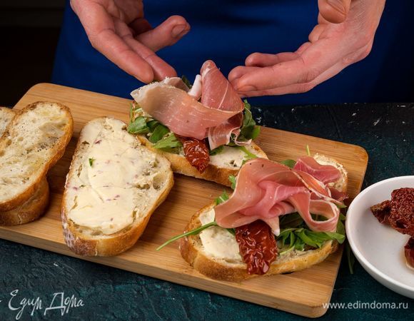 Застелите сыр листьями руколы. Сверху выложите ломтики прошутто и вяленые помидоры.