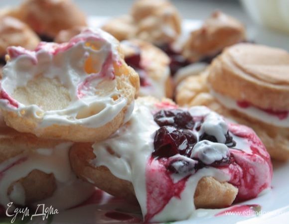 Начинаем собирать торт! Выложите на блюдо профитроли в форме венка, поместив 1–2 штуки в центр. Срежьте у профитролей верх и наполните сливочным кремом. Поверх крема выложите вишни из варенья и накройте сверху верхушками профитролей.