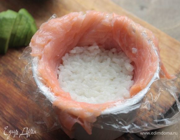 Для формирования закуски возьмите небольшие кокотницы и выстелите их пищевой пленкой. Нарежьте лосося тонкими, но большими пластами и внахлест выстелите в форму. Поверх выложите рис, чуть прижмите его и разровняйте.