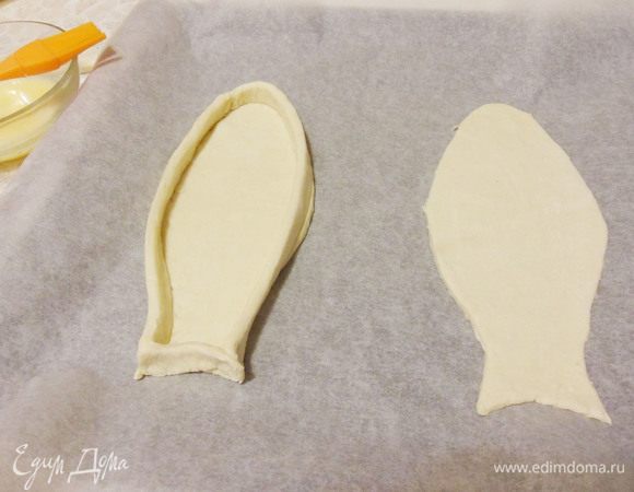 Переложить заготовки на противень застеленный бумагой для выпечки. Из обрезков теста вырезать длинные полоски, смазать их с одной стороны яйцом. Выложить полоски «бортиками» по краю рыбки, смазанной стороной вниз.