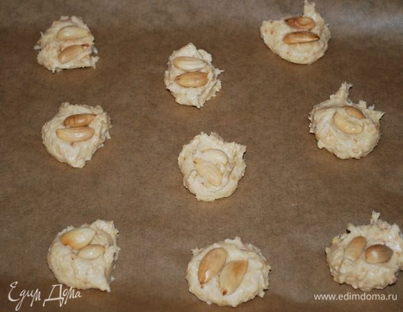 Противень застелить бумагой для выпечки, чайной ложкой выкладывать тесто так, чтобы печенья не соприкасались. Украсить печенье орехами. Выпекать в разогретой духовке при 170°С примерно 15–17 минут.