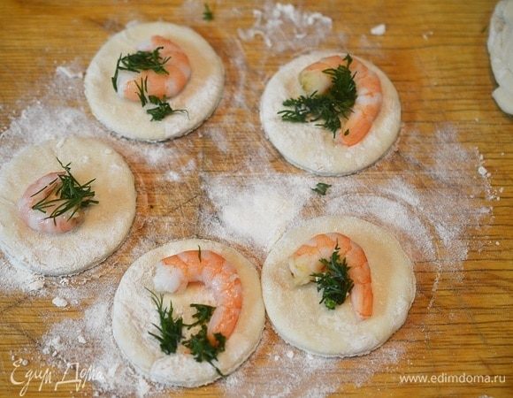 Пельменное тесто раскатайте, вырежьте кружки. В центр каждого положите немного нарезанной зелени и по одной креветке. А также, если готовите на Новый год и желаете погадать, положите по какой-нибудь добавке: монетка, перец горошком, бутон гвоздики и т.д.