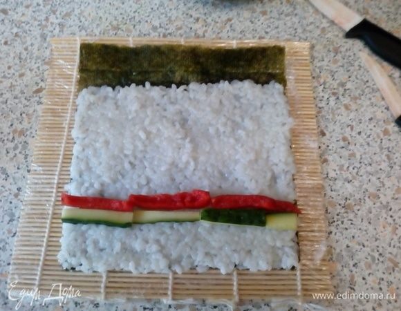 На шершавую поверхность нори выложить рис, оставив ~1–2 см листа не покрытым. Выложить бруски перца и огурца. 