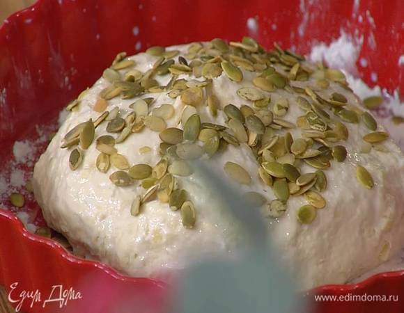 Сформировать шар из теста (оно должно слегка липнуть к рукам), выложить его в форму, так чтобы получилась круглая буханка, сделать сверху крестообразный надрез, смазать с помощью кисточки оставшимся молоком и посыпать семечками.