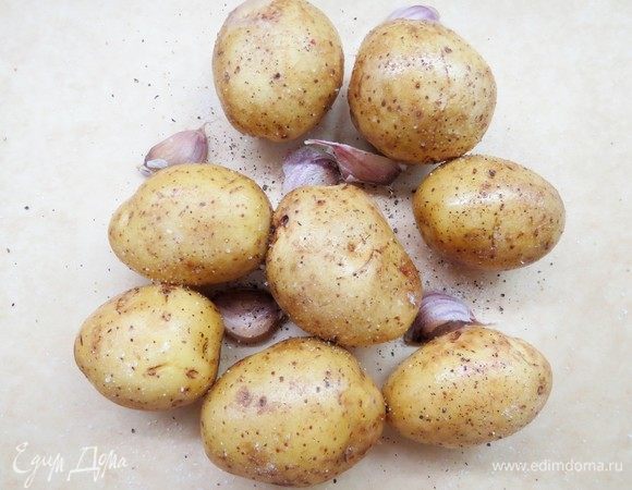 Духовку разогреть до 200°С. Картофель хорошо помыть, не чистить, добавить оливковое масло. Выложить на противень лист пергамента, визуально поделить его пополам. Выложить смазанный маслом картофель на половину пергамента, между картофелем разложить нечищенные дольки чеснока. Посолить и поперчить.