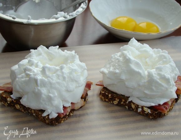 Сверху на бекон выложить взбитые белки в виде «шапочки», не забыв сделать в центре ямочку для желтков. 
Поставить бутерброды в разогретую до 180°С духовку минут на 10.
