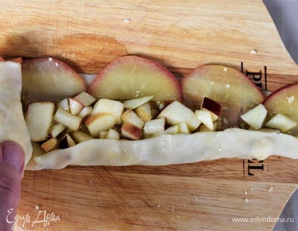 Делаем розы. Полоску тесто смазываем маслом, посыпаем тонким слоем орехов и яблок. Полукольца яблок в ряд кладем по верхнему краю в длину, чтобы 0,5 см яблока с кожицой выступала над тестом. Сворачиваем штрудель трубочкой, предварительно 1 см нижнего края завернув. Вертикально ставим на лист с бумагой для запекания полукольцами яблок наверх. 