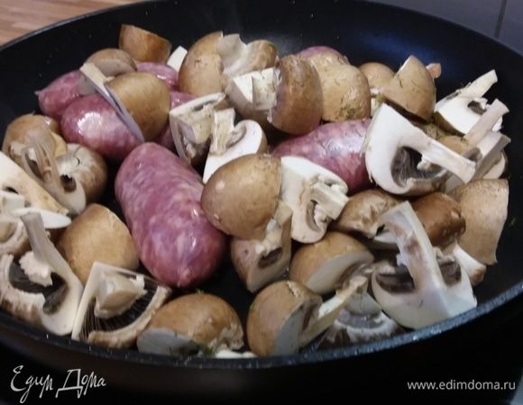 Выложить к колбаскам шампиньоны, нарезанные на половинки или мельче, в зависимости от их размера.